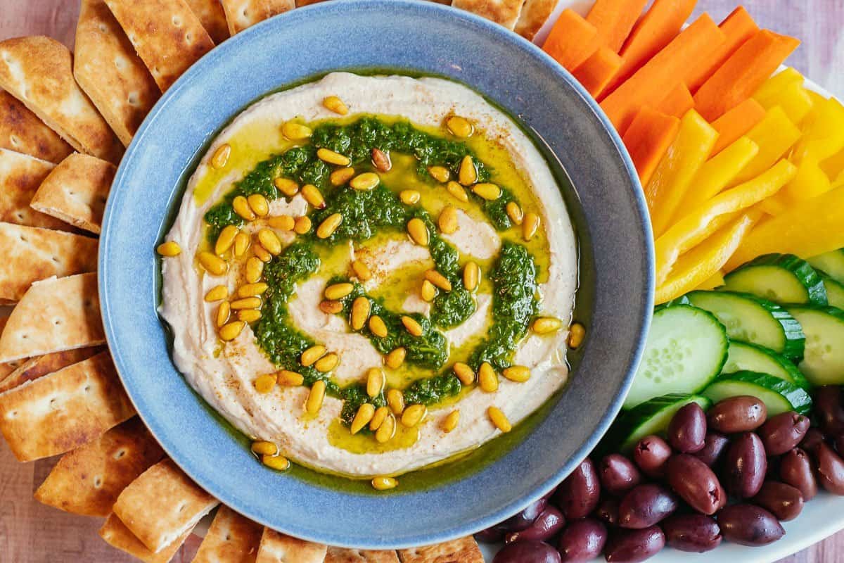 white bean hummus in a bowl topped with herb salsa and spiced pine nuts, surrounded by carrots, cucumbers, olives, and pita bread