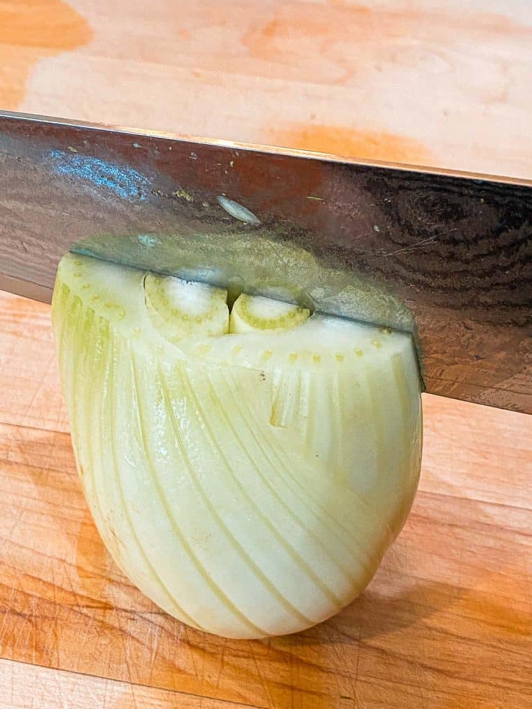 fennel bulb being sliced in half from top to bottom with a knife, depicting how to cut fennel to use in a vegetable side dish.
