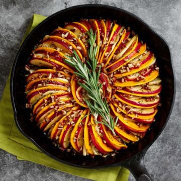 Butternut squash and apple tian with fennel and orange is pictured in a cast iron skillet, garnished with rosemary.