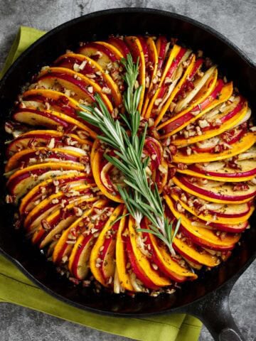 Butternut squash and apple tian with fennel and orange is pictured in a cast iron skillet, garnished with rosemary.