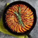 Butternut squash and apple tian with fennel and orange is pictured in a cast iron skillet, garnished with rosemary.