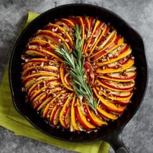 Butternut squash and apple tian with fennel and orange is pictured in a cast iron skillet, garnished with rosemary.