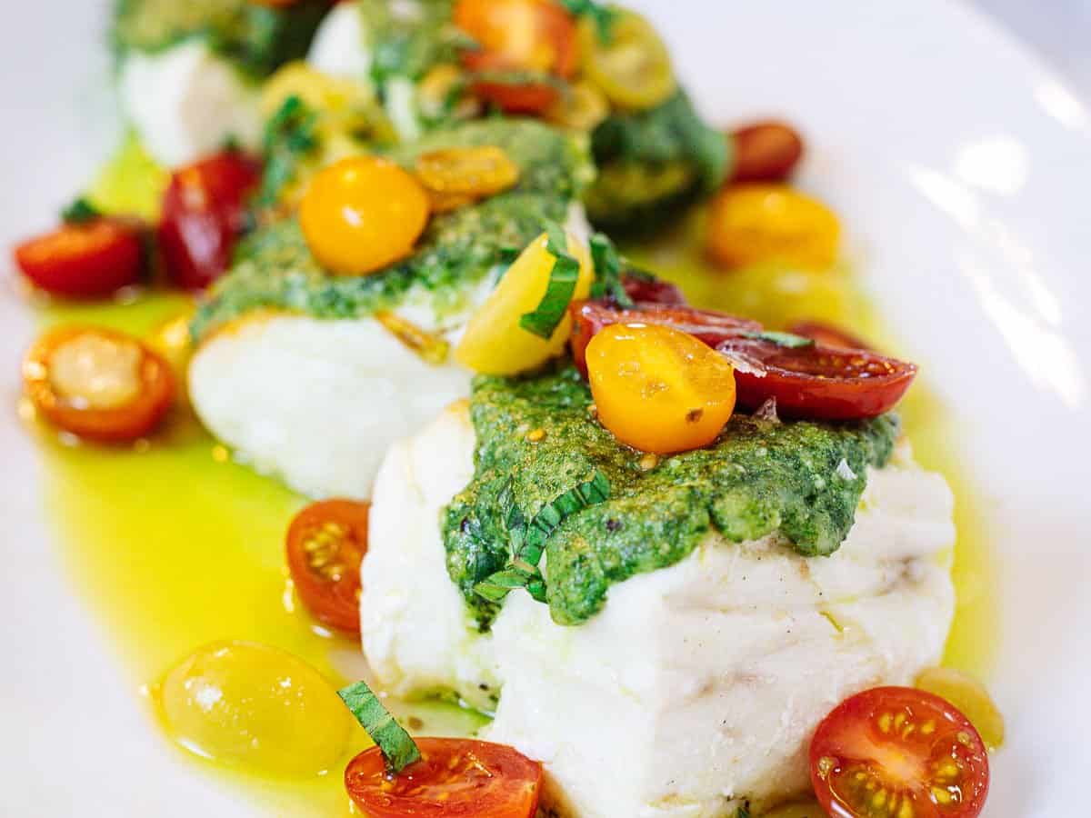 Pieces of sear-roasted halibut topped with macadamia nut pesto and tomatoes on a white plate