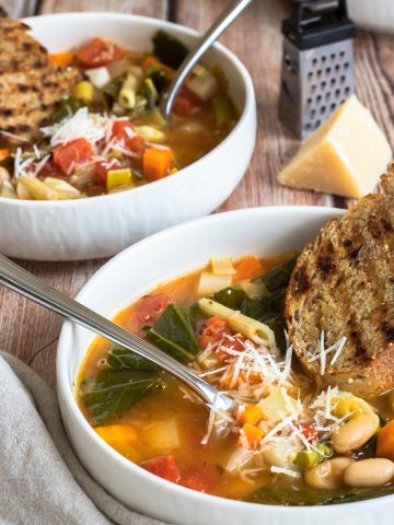 two white bowls filled with minestrone soup made with leeks, asparagus, and kohlrabi. Topped with grilled whole grain bread and parmesan cheese.