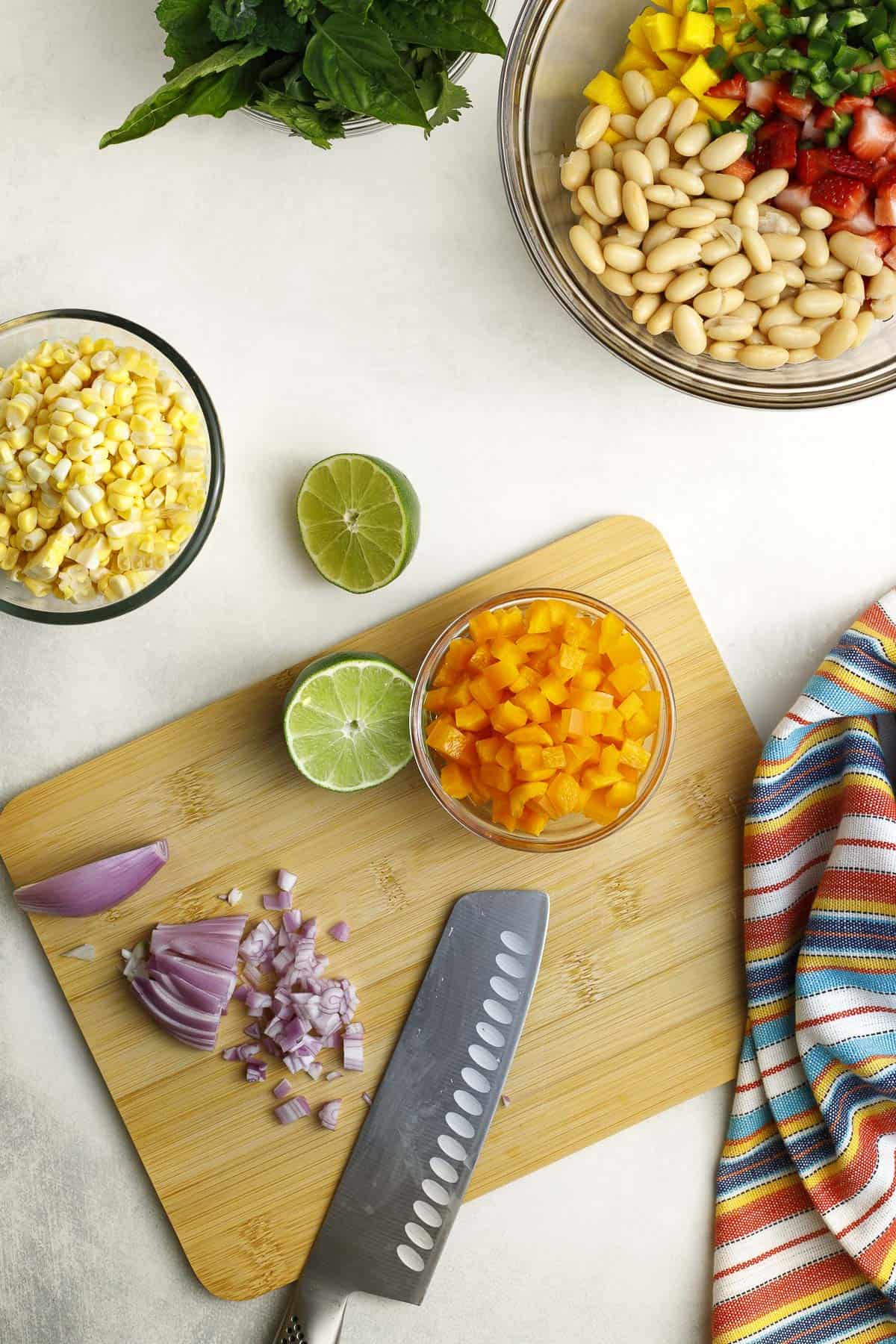 chopped shallots and a knife on a cutting board surrounded by diced bell pepper in a bowl, corn kernels in a bowl, and a lime.
