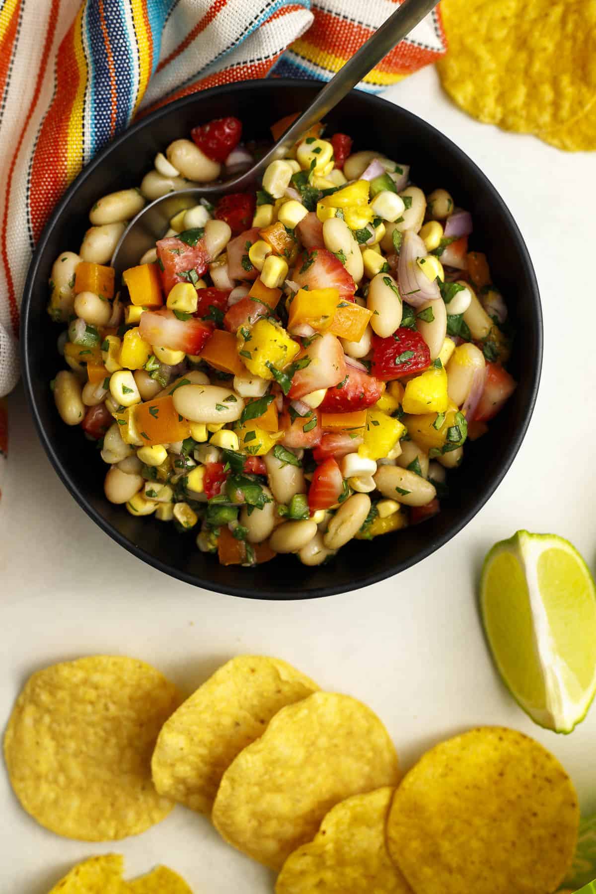 Cowgirl caviar with mango, strawberries, white beans, and corn in a bowl with chips and lime wedges on the side.