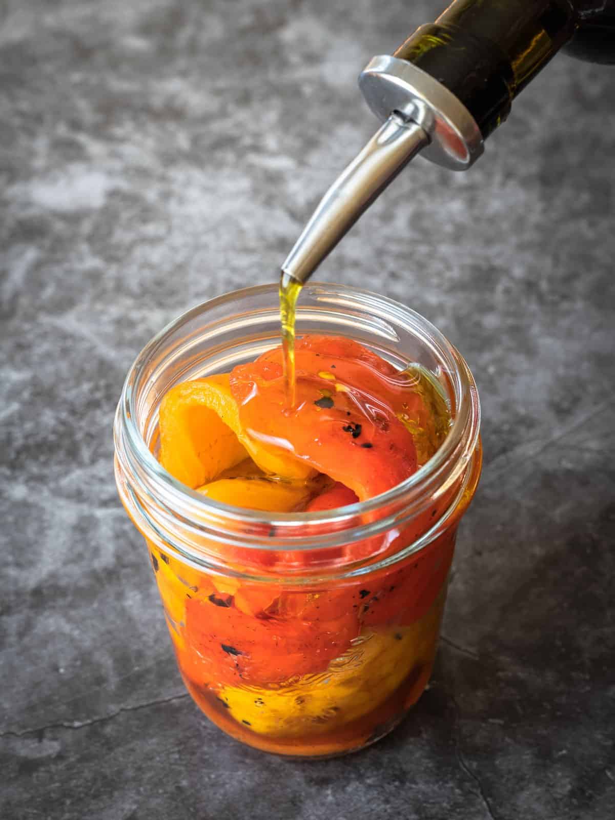 Olive oil being poured over roasted bell peppers in a glass jar