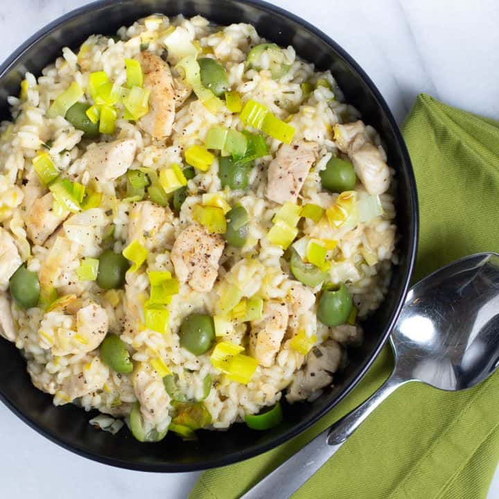 Risotto with chunks of chicken, green olives, and leeks in a black bowl with a napkin and a spoon