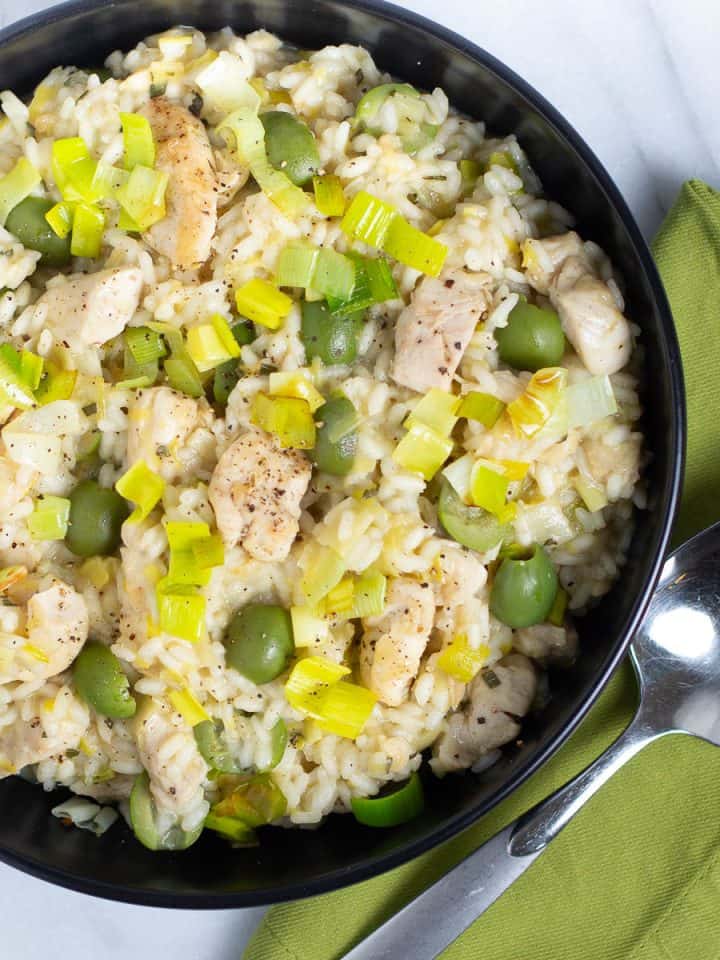 Risotto with chunks of chicken, green olives, and leeks in a black bowl with a napkin and a spoon