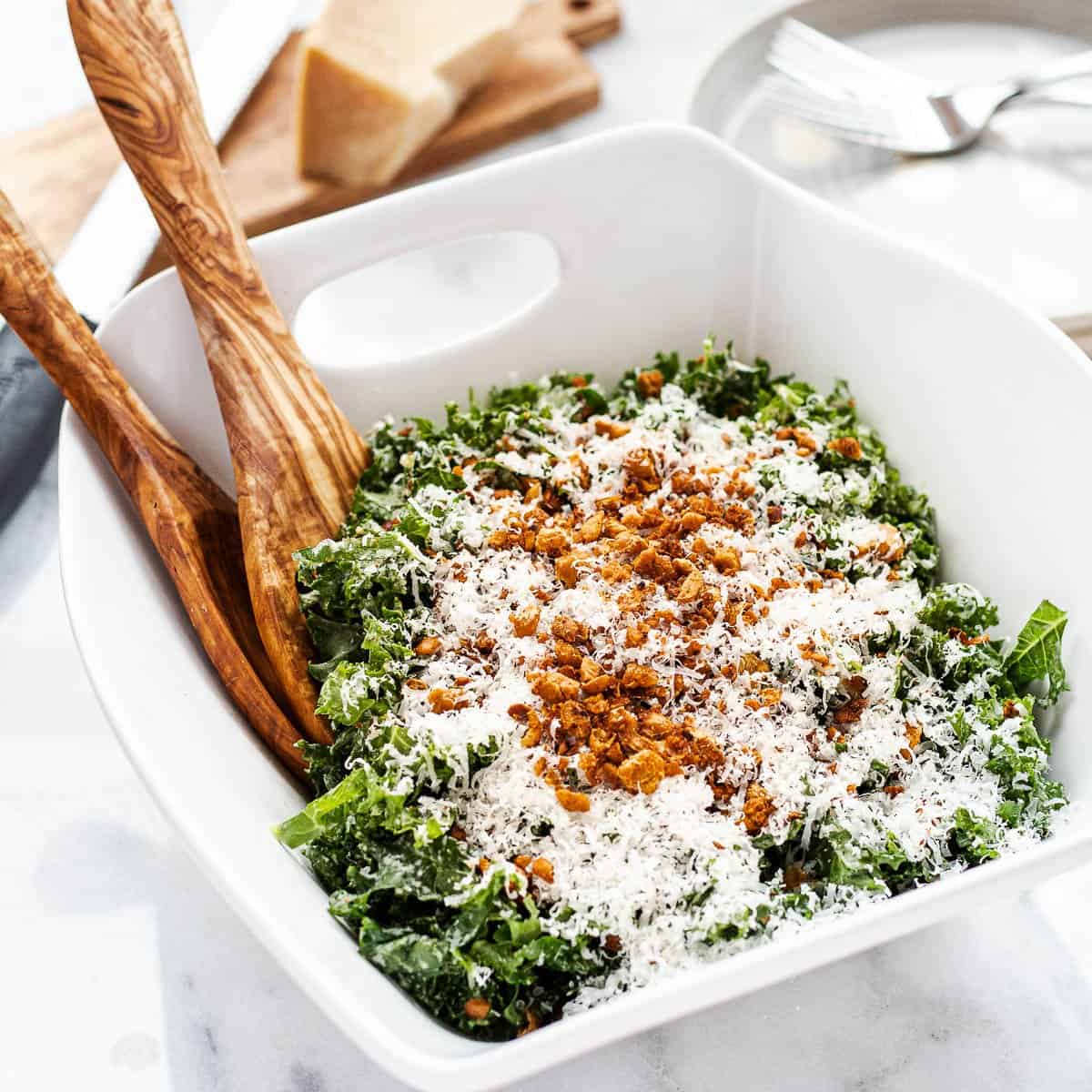 Kale tahini Caesar salad in a serving bowl topped with crispy harissa chickpeas and shredded Parmesan. Accompanied by salad tongs.