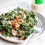 tahini kale Caesar salad on a plate with a fork. Topped with crispy harissa roasted chickpeas and shredded parmesan. Accompanied by a fork and a jar of dressing in the background.