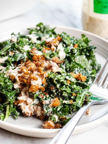 tahini kale Caesar salad on a plate with a fork. Topped with crispy harissa roasted chickpeas and shredded parmesan. Accompanied by a fork and a jar of dressing in the background.