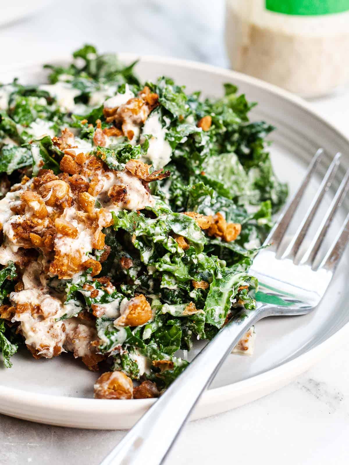 tahini kale Caesar salad on a plate with a fork. Topped with crispy harissa roasted chickpeas and shredded parmesan. Accompanied by a fork.