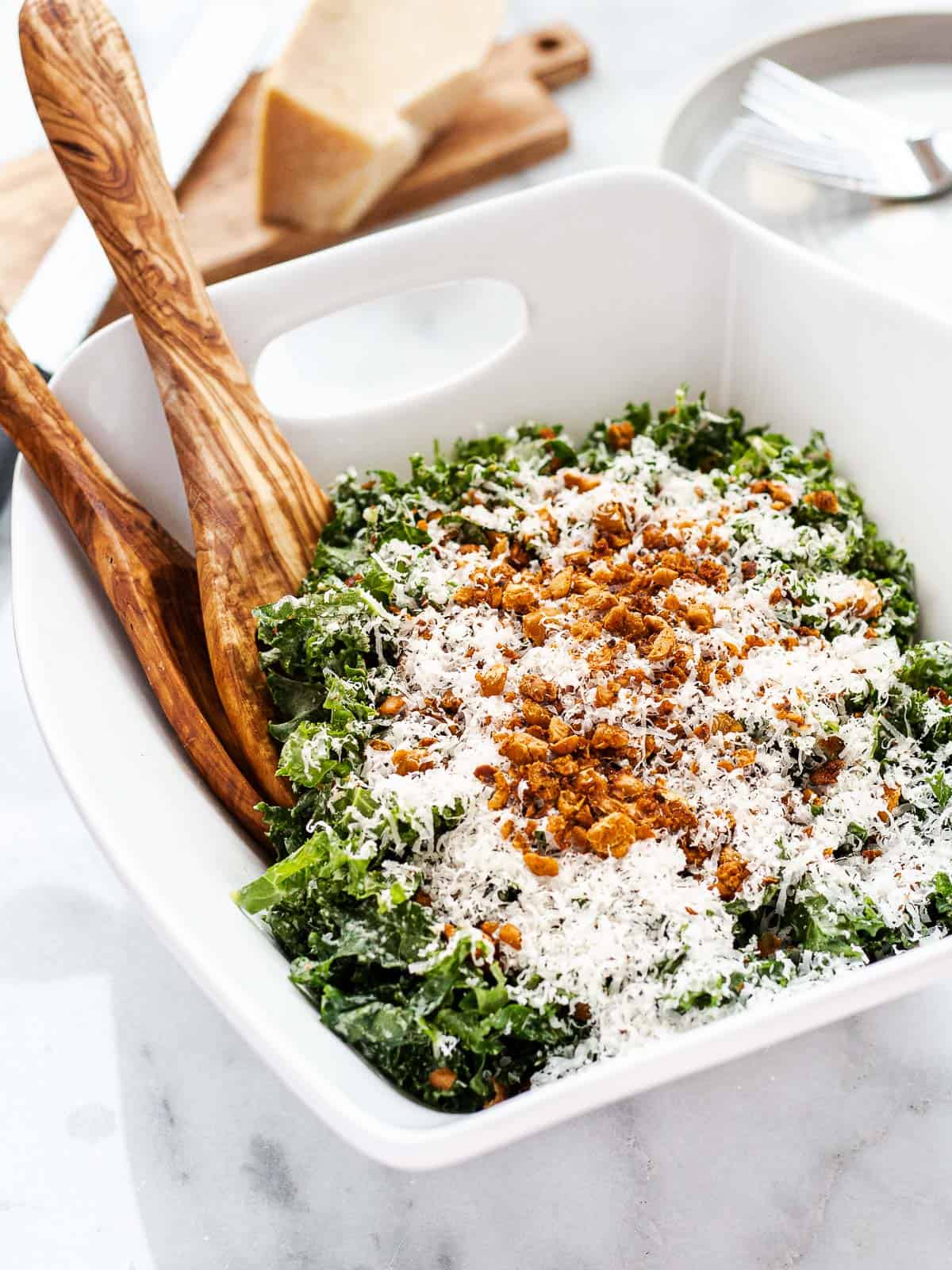 Kale tahini Caesar salad in a serving bowl topped with crispy harissa chickpeas and shredded Parmesan. Accompanied by salad tongs.