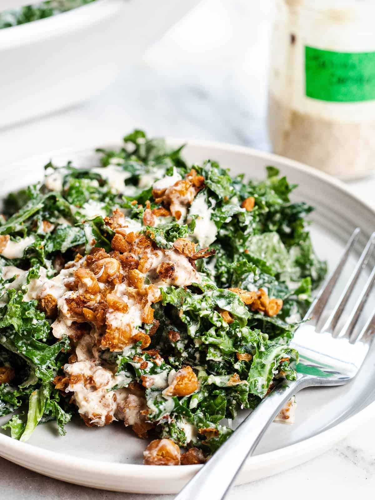 tahini kale Caesar salad on a plate with a fork. Topped with crispy harissa roasted chickpeas and shredded parmesan. Accompanied by a fork and a jar of dressing in the background.