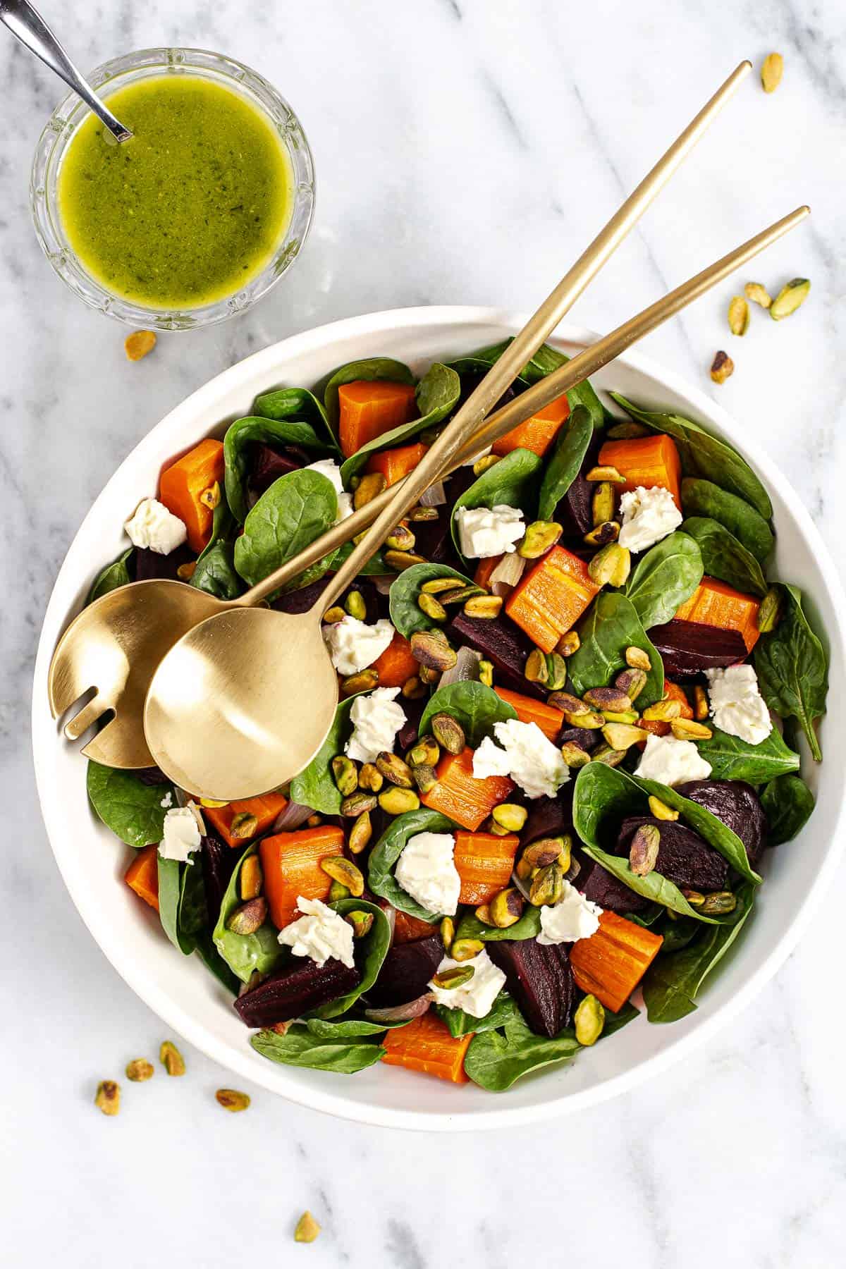 Roasted beet and carrot salad on a bed of spinach and topped with feta and pistachios in a bowl with salad tossers. A bowl of tarragon vinaigrette sits next to it. 