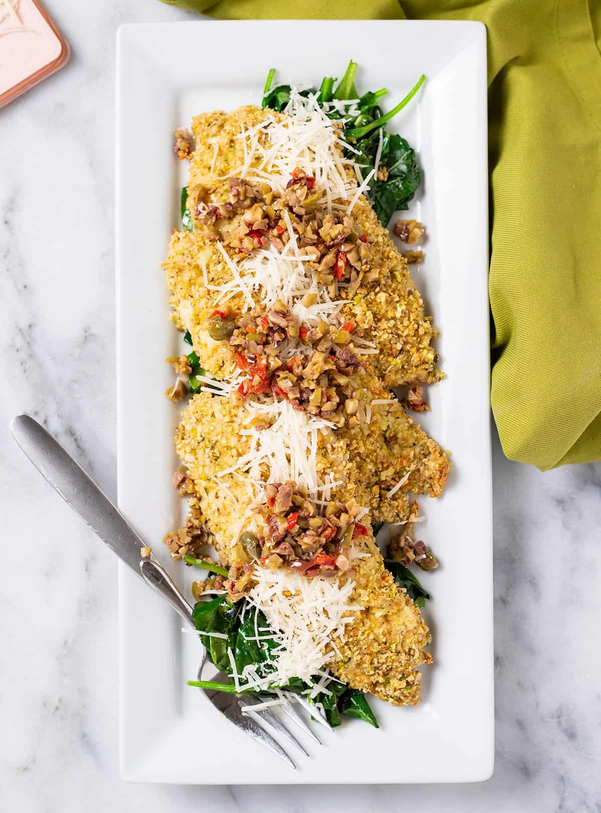 Almond and asiago crusted rockfish Mediterranean diet dinner. Fish is on a plate topped with olive tapenade and shredded cheese. Accompanied by a serving fork and a napkin.