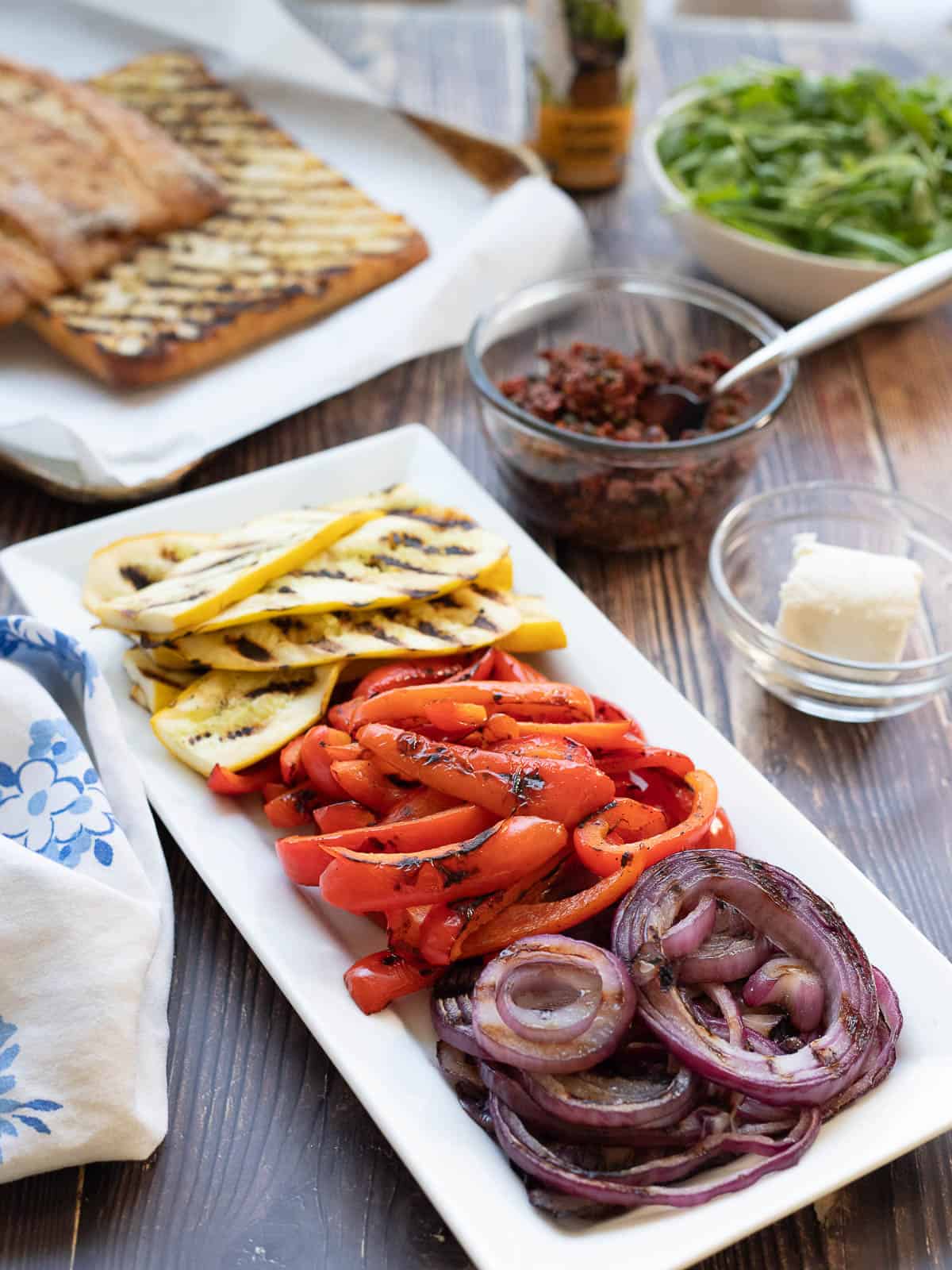 All the fixings for grilled vegetable sandwich: grilled summer squash, bell peppers, and onions on a plate, goat cheese in a bowl, sundried tomato and olive tapenade in a bowl, ciabatta cut in half and grilled, a bowl of arugula, and balsamic drizzle.