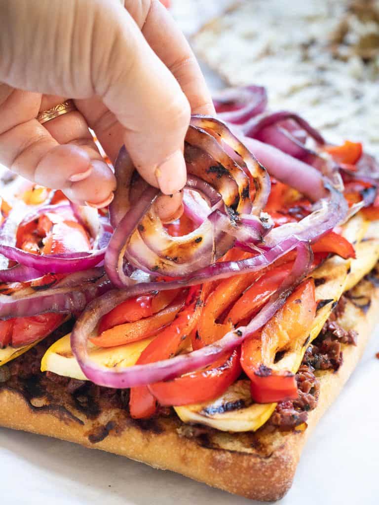 Grilled onions being layered onto ciabatta layered with sundried tomato and olive tapenade, grilled summer squash and bell peppers.