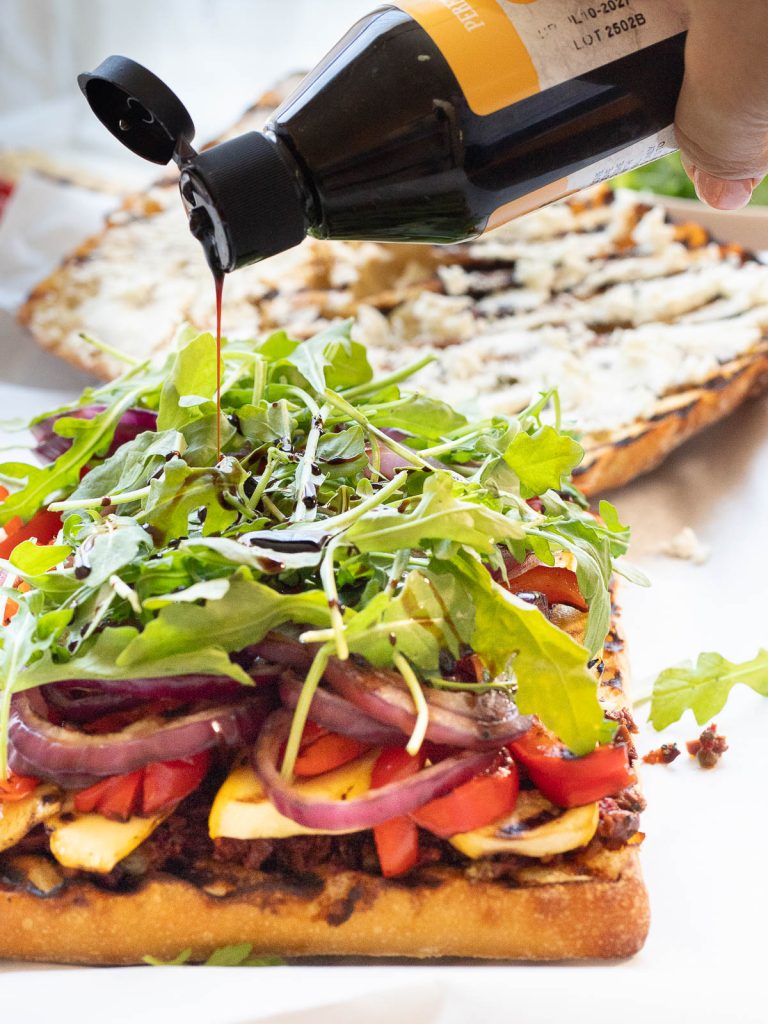 Balsamic sauce being drizzled on grilled ciabatta layered with grilled vegetables, sundried tomato and olive tapenade, and arugula. The top of the ciabatta is in the background spread with goat cheese.