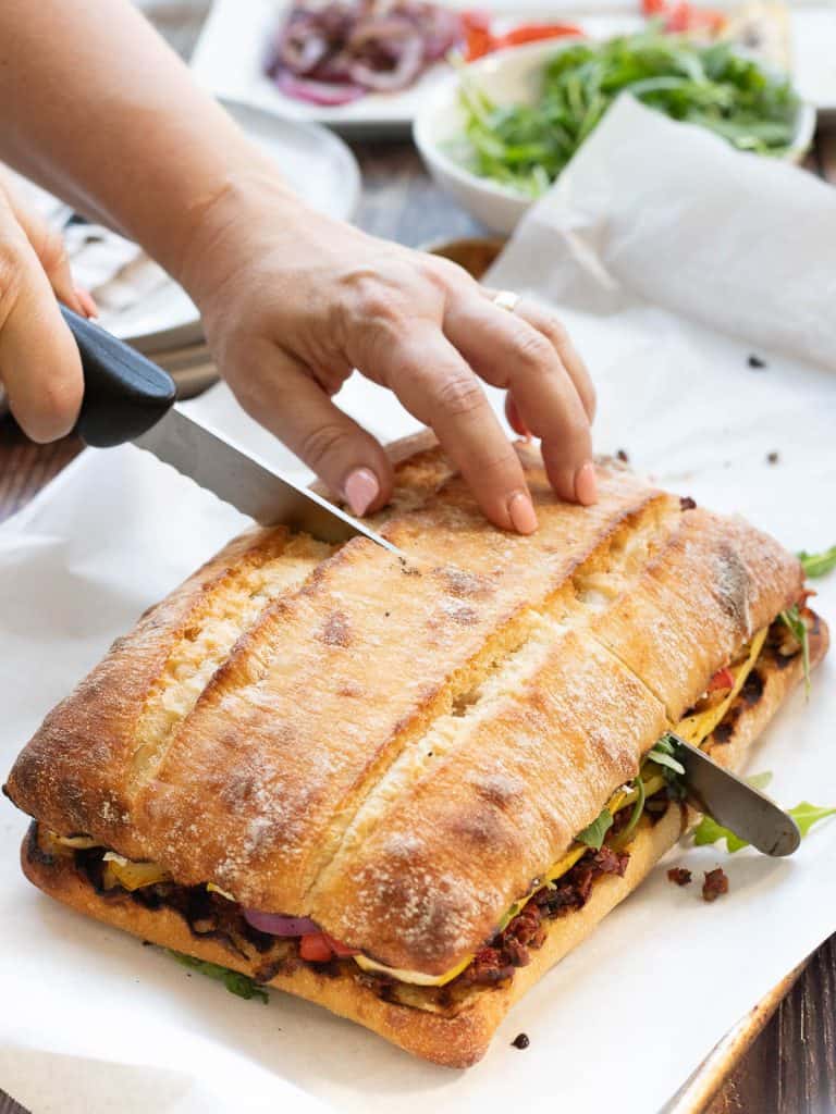 Sandwich on ciabatta being cut with a bread knife. Grilled vegetables and a bowl of arugula are in the background.