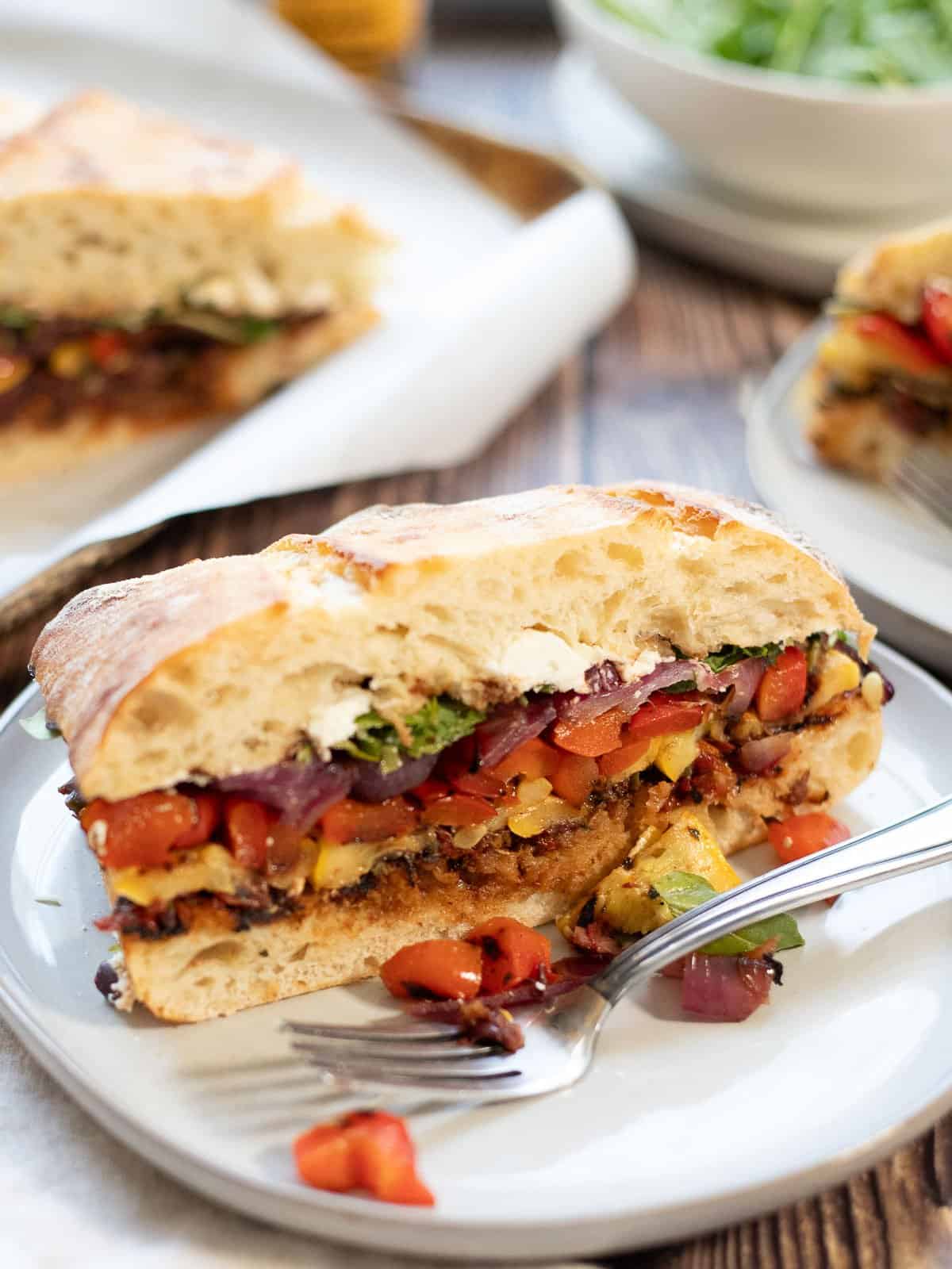 Grilled vegetable sandwich on a plate with a fork. More sandwiches in the background, along with a bowl of arugula.