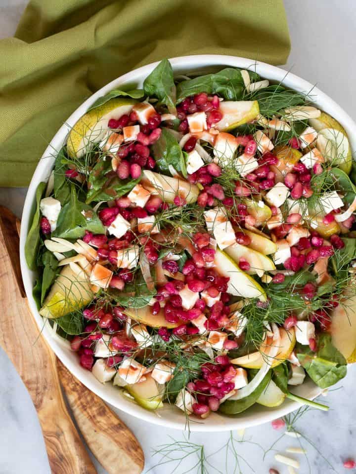 Pear and fennel salad in a bowl with salad tongs and a napkin. Garnished with toasted almonds, feta, and pomegranate arils.