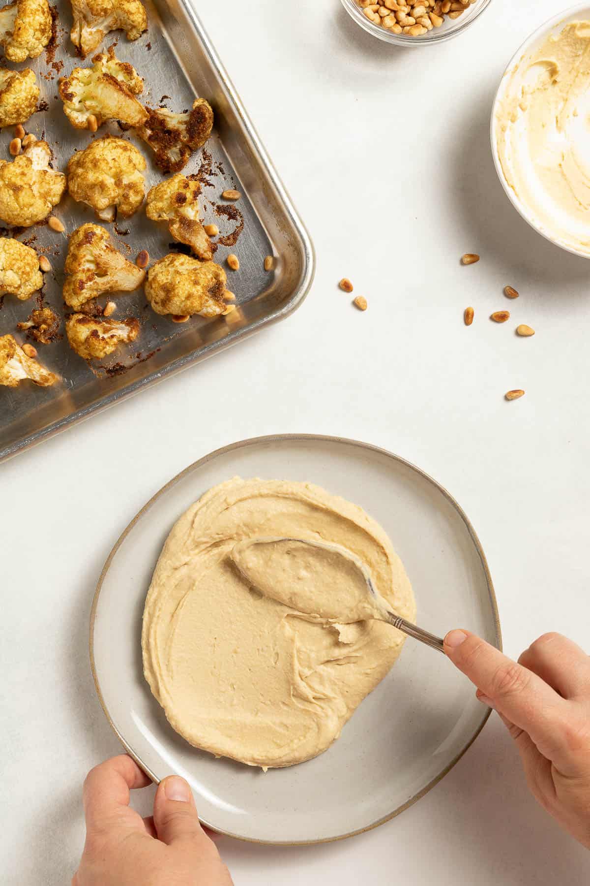 Garlic hummus being spread on a plate with a spoon. A baking sheet with roasted cauliflower sits next to it, along with a bowl of toasted pine nuts.