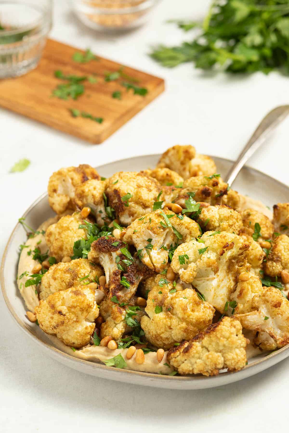 Coriander roasted cauliflower is served over a platter of hummus with a spoon. It is garnished with toasted pine nuts and parsley. Bowls filled with garnishes are in the background.