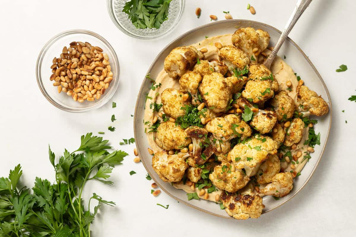 Cauliflower roasted with coriander and cumin is served over a platter of hummus with a spoon. It is garnished with toasted pine nuts and parsley. Accompanied by a small bowl of toasted pine nuts and a small bowl of chopped parsley.