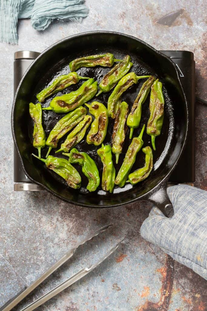 A cast iron pan filled with blistered shishito peppers sits on a portable burner. The handle is being held by someone wearing an oven mitt.