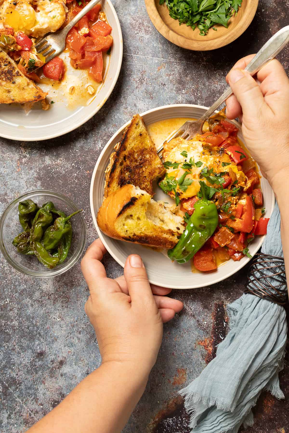 A serving of shakshuka on a plate with grilled bread and a blistered shishito pepper as garnish. A hand is holding the plate and another is holding a fork and digging in to the poached egg in the shakshuka. A bowl of blistered shishito peppers sits next to the plate.