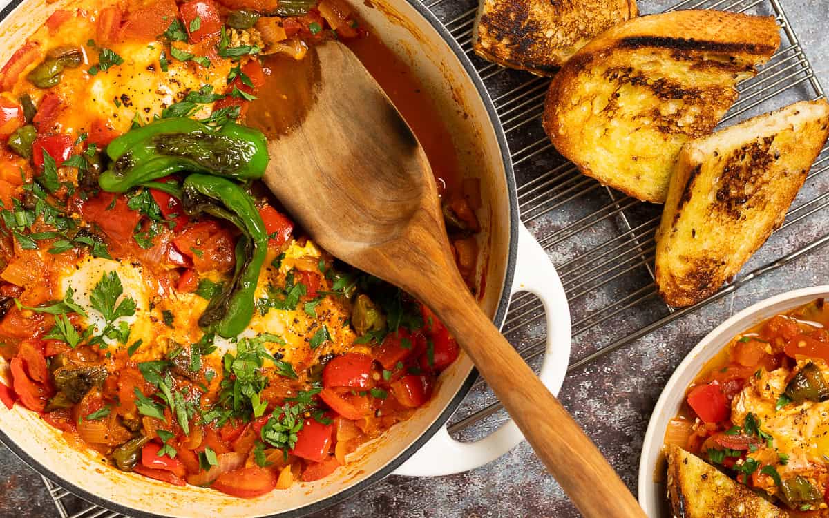 A close up of a pot of shakshuka with a serving utensil. Bread is served on the side.