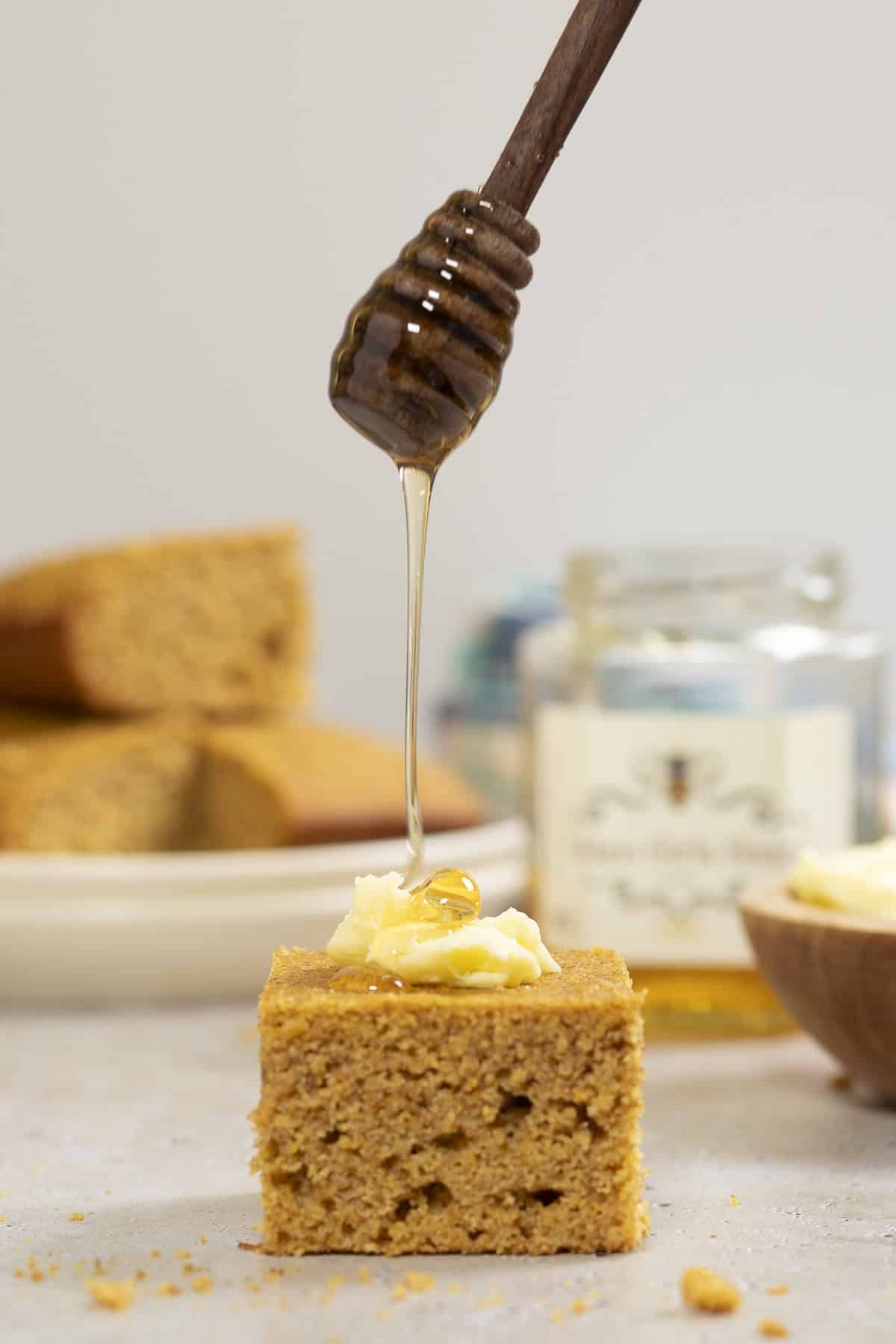 Honey is being drizzled on a piece of cornbread that is topped with a bit of butter. A plate full of whole wheat cornbread, a bowl of butter, and a pot of honey sit behind it.