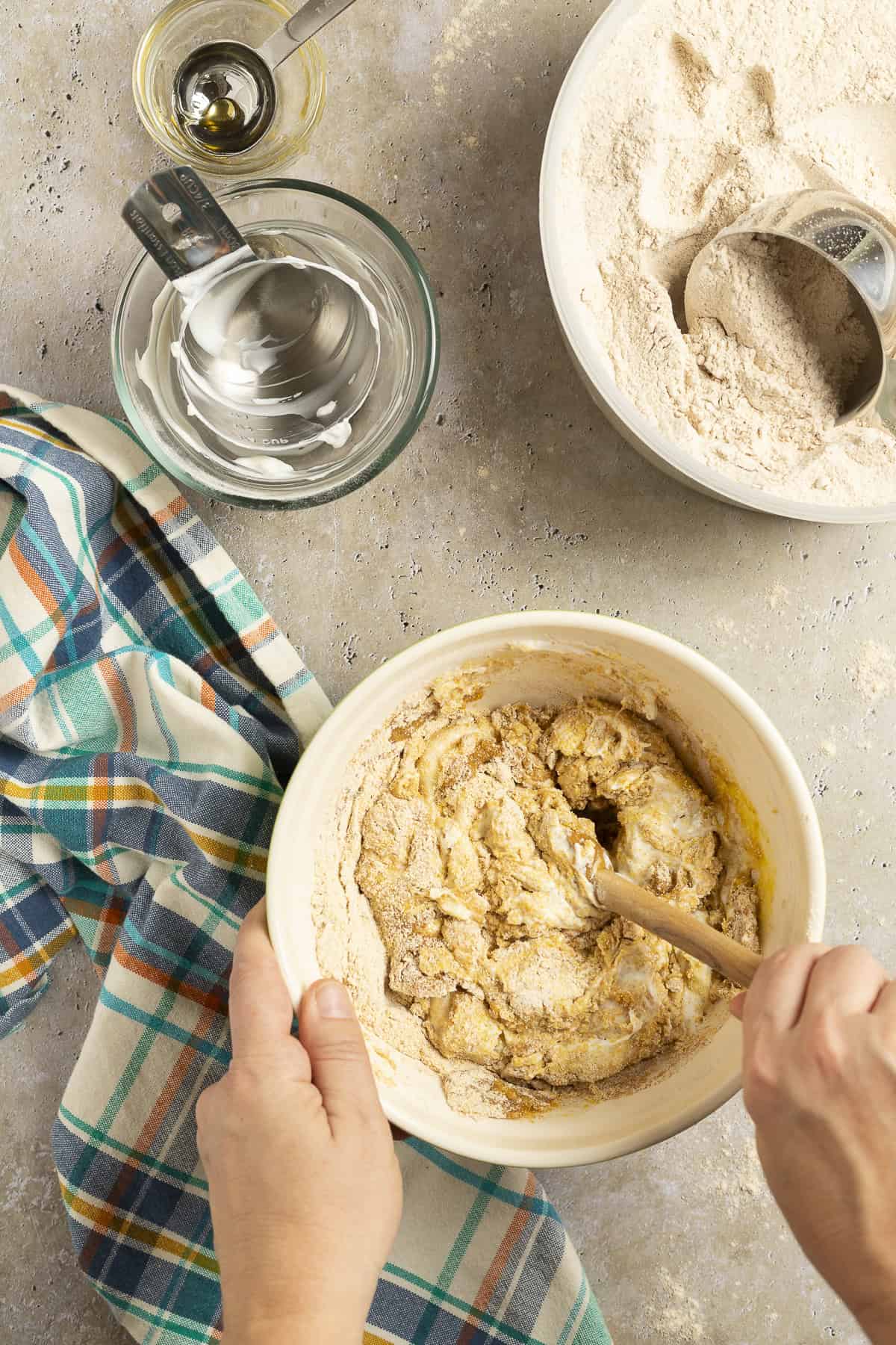 A bowl of cornbread batter is being mixed with a spoon. A container of whole wheat flour sits nearby with a cup measure in it.