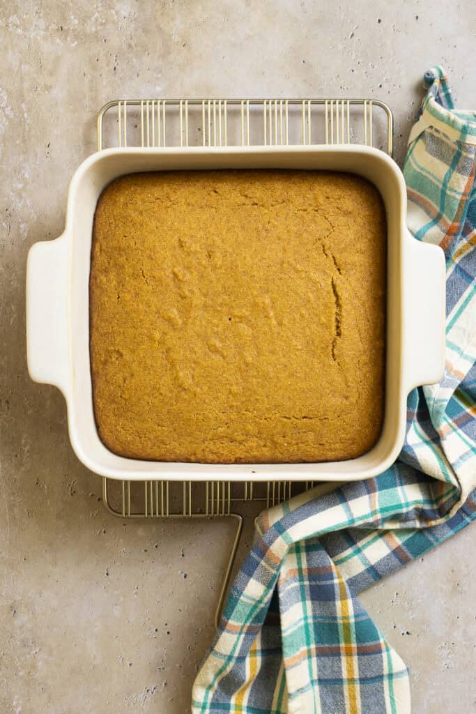 A pan of cornbread sits on a cooling rack with a dishtowel.