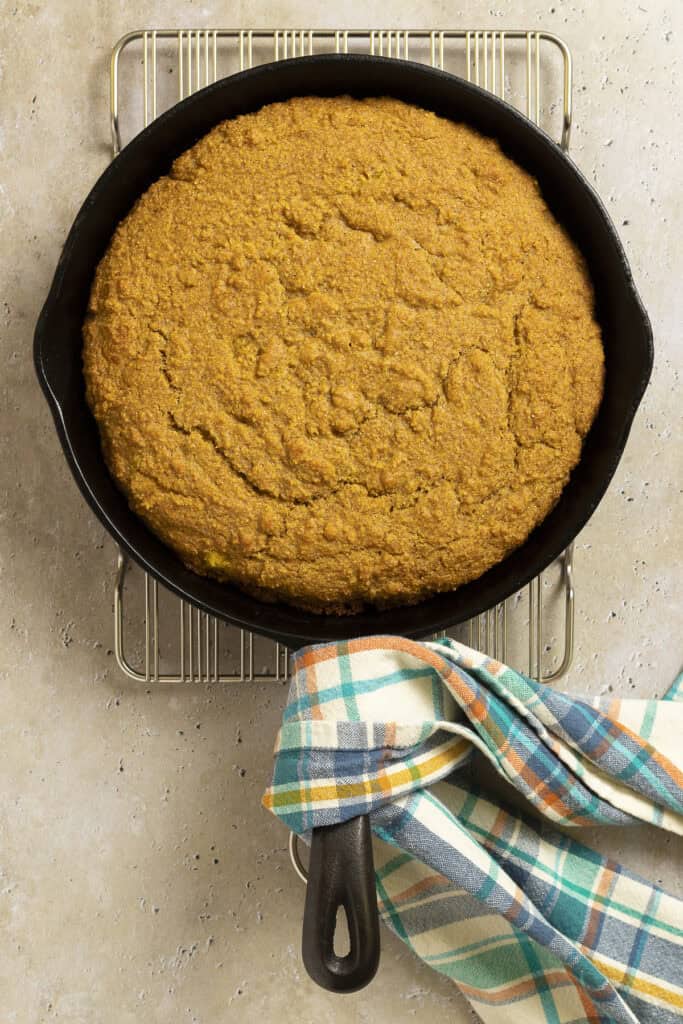 A pan of cornbread cooked in a skillet sits on a cooling rack with a dish towel.