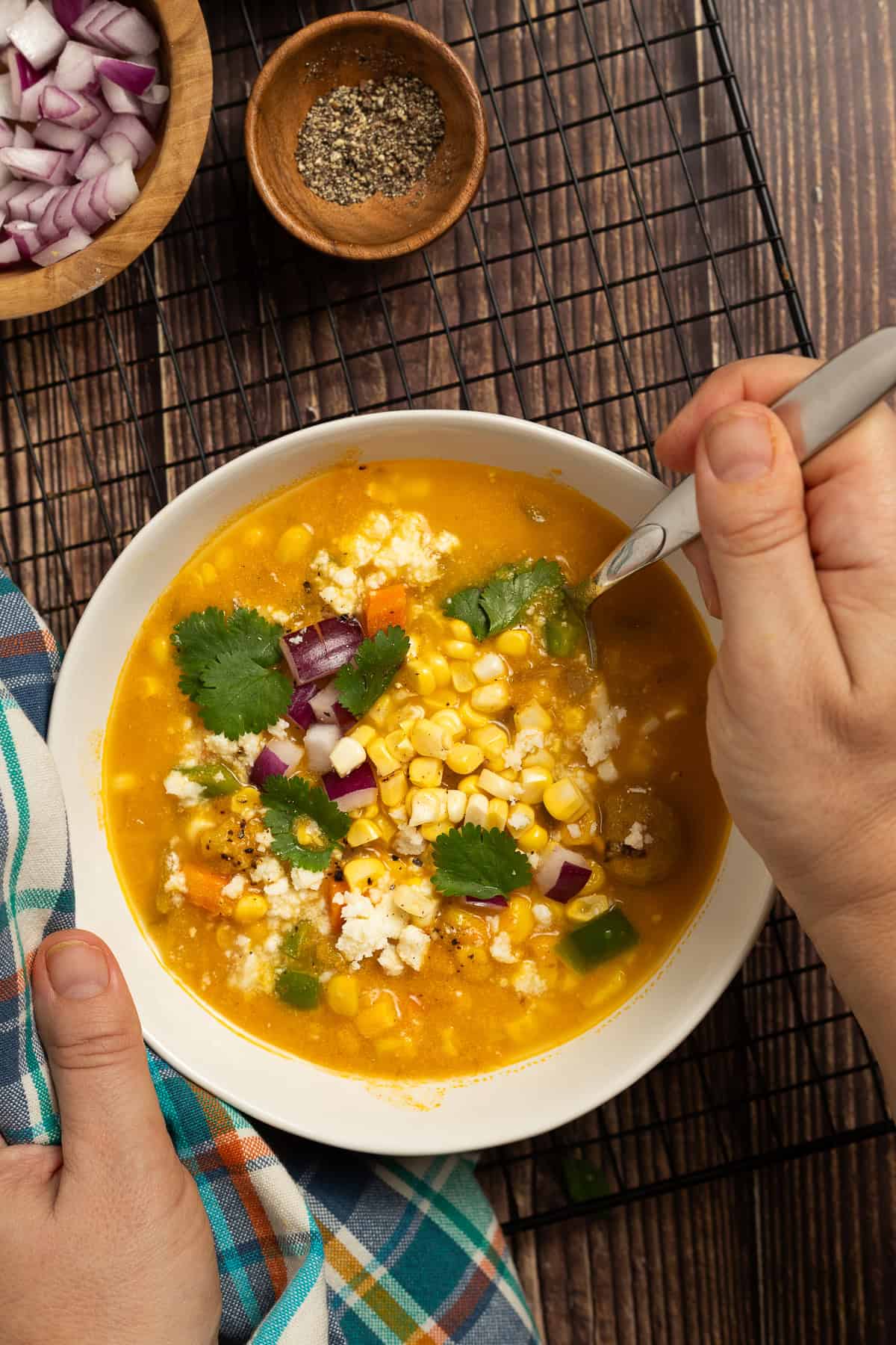 Two hands holding a bowl of pumpkin corn chowder garnished with corn, red onion, cilantro, and queso fresco.