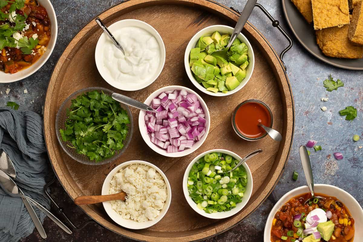 Toppings for smoky vegetarian chili with beans: plain Greek yogurt, chopped avocado, hot sauce, sliced green onions, queso fresco, chopped red onion, and cilantro leaves.