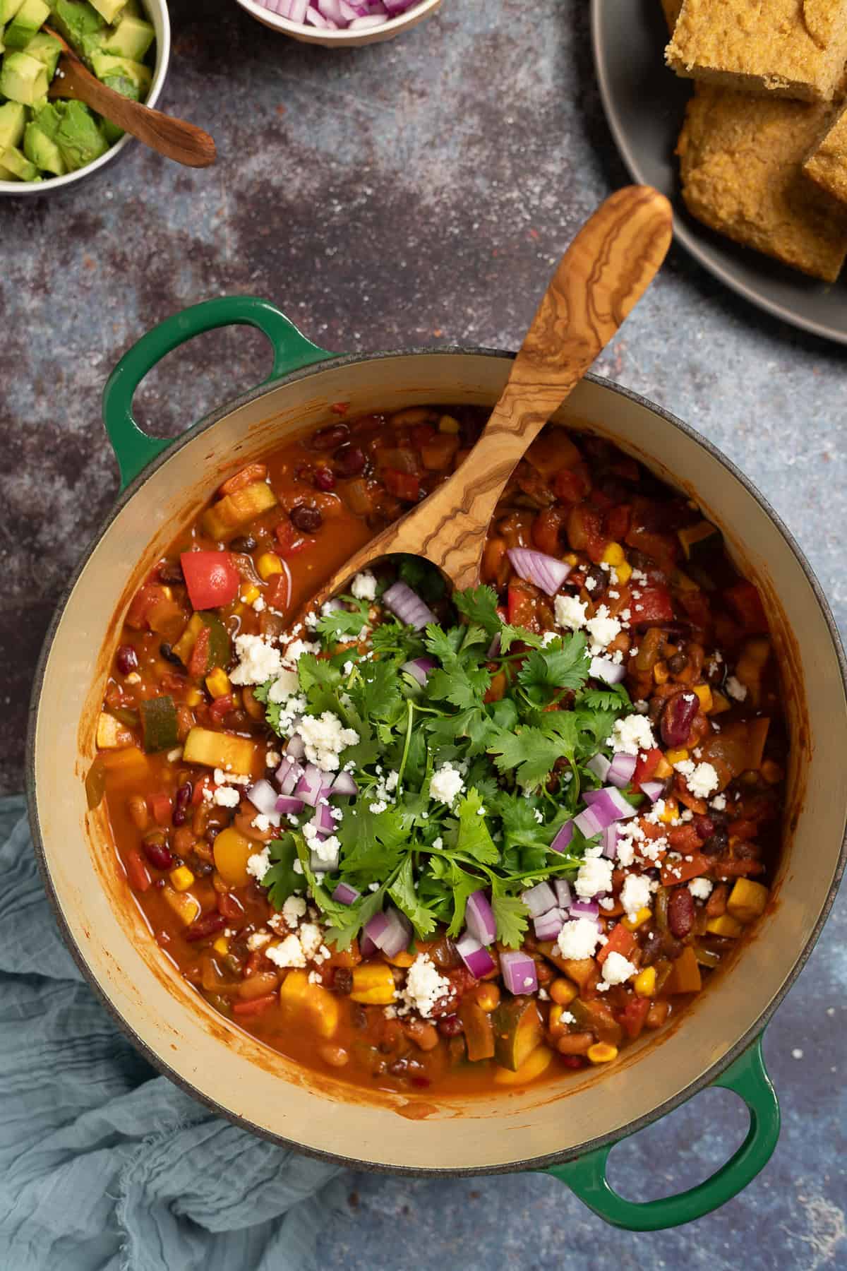 A big pot of chili sits on a countertop with a spoon. It is topped with cheese, red onions, and cheese. Cornbread sits on a plate to the side. 