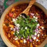 A pot of vegetarian chili with a spoon. Topped with cheese, red onion, and cilantro.