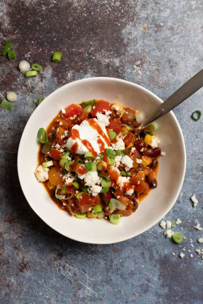 A bowl of vegetarian chili is shown topped with plain yogurt, hot sauce, and sliced green onions.