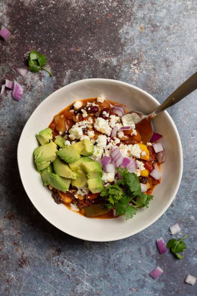 A bowl of vegetable chili with beans is shown topped with avocado, red onion, cilantro, and queso fresco.