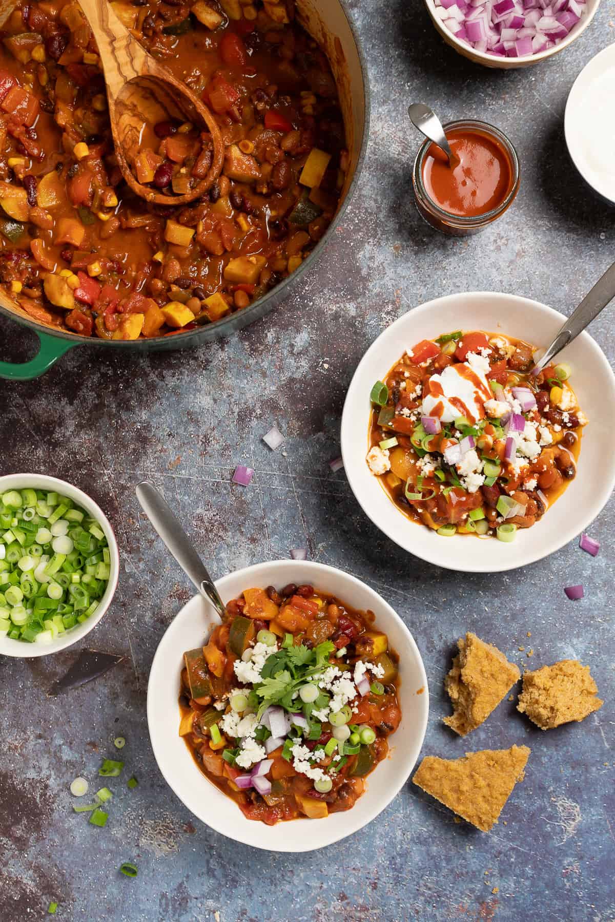 Two bowls of vegetarian chili sit next to a pot of chili. Toppings sit to the side, such as green onions, red onions, cheese, and hot sauce. Cornbread sits next to the bowls.