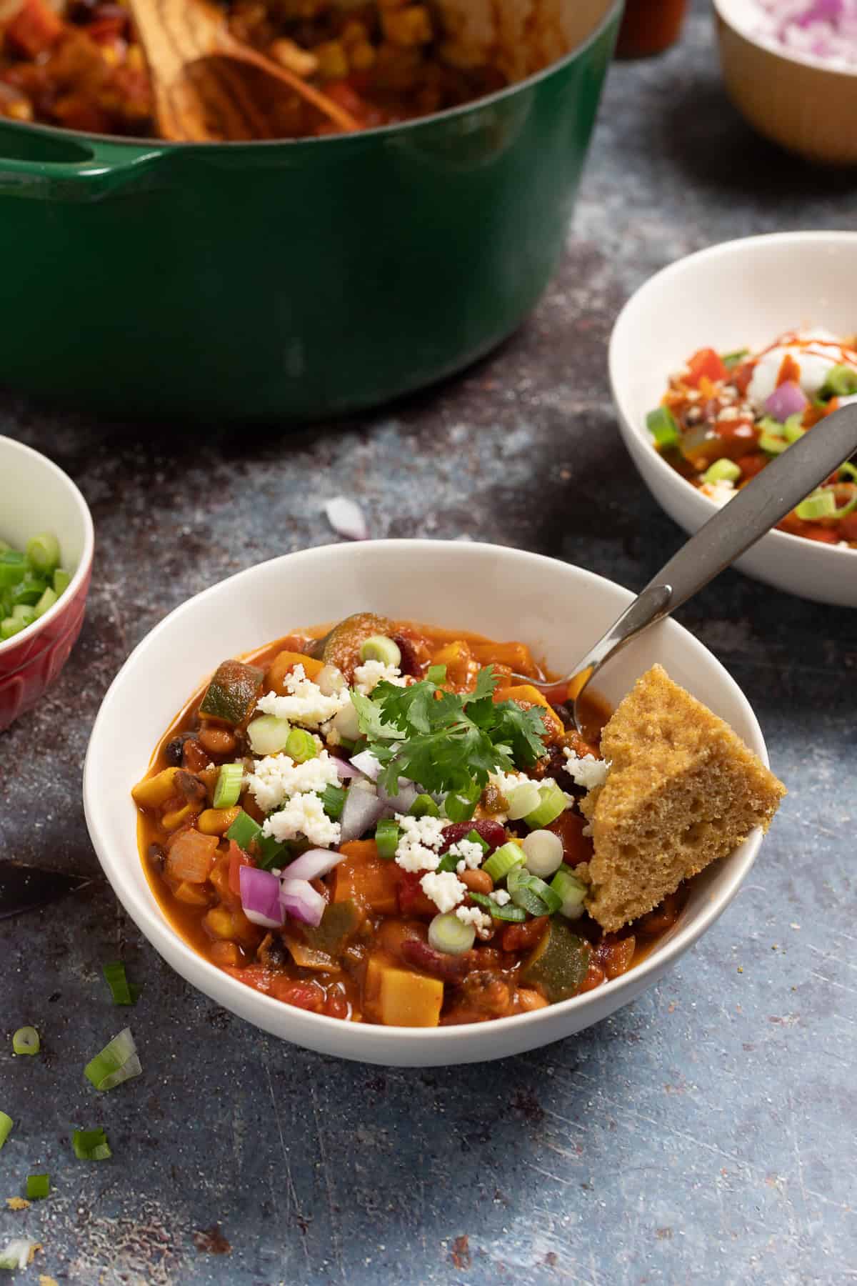 Vegetable chili in a bowl with a spoon. Topped with queso fresco, cilantro, red onion, and cornbread. 