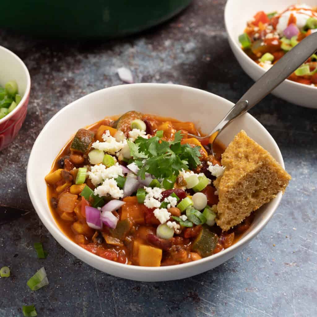 A bowl of smoky vegetarian chili with beans in a bowl topped with queso fresco, cilantro, red onions, and cornbread.