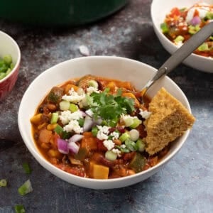 A bowl of smoky vegetarian chili with beans in a bowl topped with queso fresco, cilantro, red onions, and cornbread.