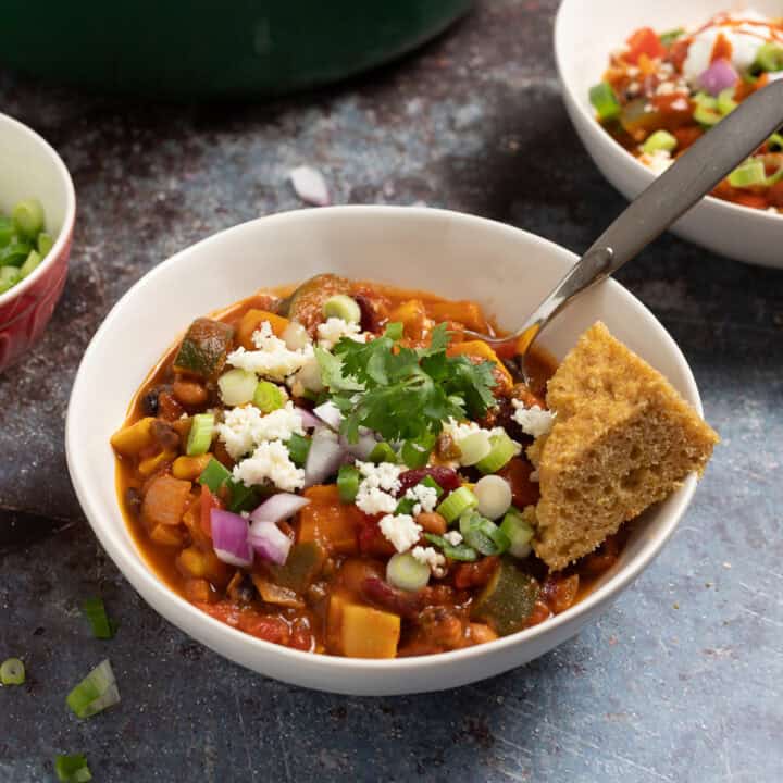 A bowl of smoky vegetarian chili with beans in a bowl topped with queso fresco, cilantro, red onions, and cornbread.