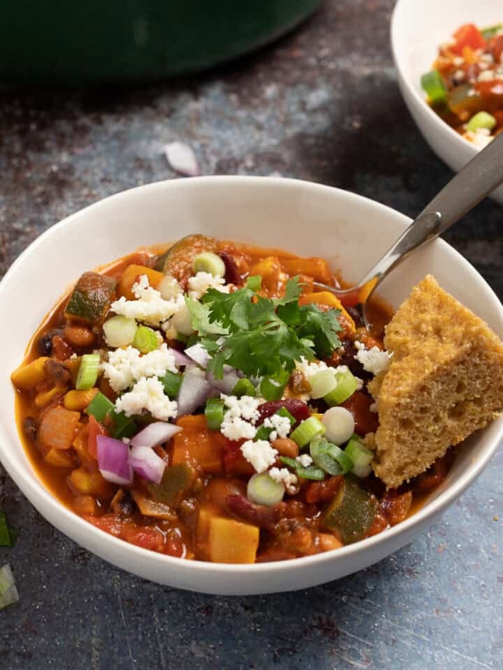 A bowl of smoky vegetarian chili with beans in a bowl topped with queso fresco, cilantro, red onions, and cornbread.