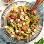 A bowl of walnut pesto pasta salad with roasted vegetables sits on a table with a napkin and a serving spoon. A bowl of walnuts sits off to the side.